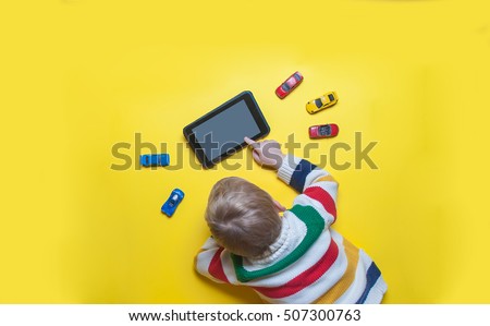 Boy using tablet and laptop while playing with toy cars on a carpet at home. Modern family. Tablet pc hero header image. Boy using digital tablet while lying on yellow floor.