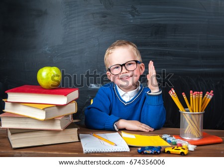 Happy cute clever boy is sitting at a desk in a glasses with raising hand. Child is ready to answer with a blackboard on a background. First time to school. Back to school. Apple and books on desk