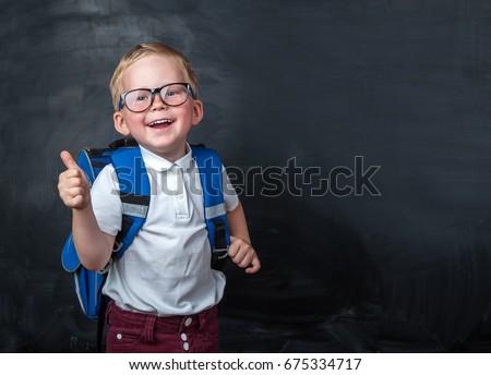 Happy smiling boy in glasses with thumb up is going to school for the first time. Child with school bag and book. Kid indoors of the class room with blackboard ona background. Back to school.