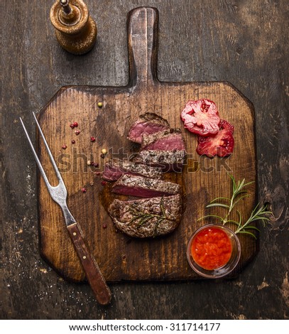 roasted  ribeye steak sliced ??on a cutting board with a fork, red sauce, peppers and tomatoes on rustic wooden background, top view