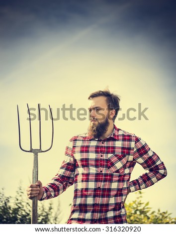 Portrait of young bearded farmer in red checkered shirt with old pitchfork on sky  nature background, toned