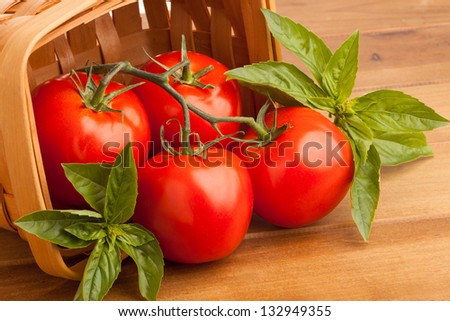 Surrounded by sweet basil, four red juicy vine ripened tomatoes fall out of a woven basket. A wooden cutting board provides the final resting place for the tomatoes before they are sliced open.