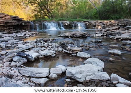 A waterfall supplies a rocky stream with an ample flow of springtime water. Along the stream, pools of water are formed reflecting the soft colors of the morning sunlit trees.