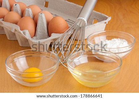 Separated cracked eggs in glass bowls, flour in a glass bowl, a silver whisk, and carton of eggs all on a cutting board.