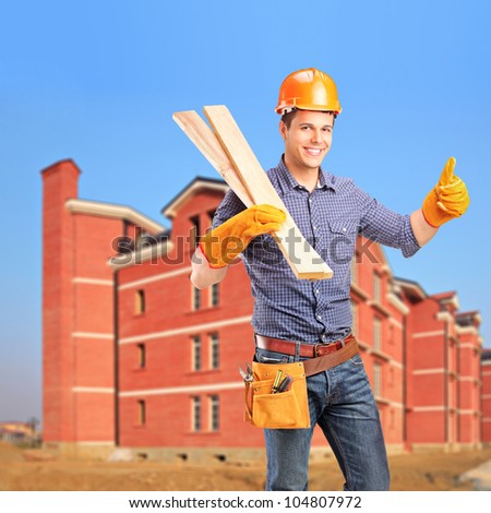 A smiling carpenter holding sills and giving thumb up with a newly built apartment block in the background