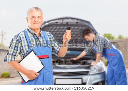 Mature mechanic in uniform holding a car key and another mechanic performing a car check in the background