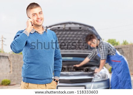 Smiling careless guy talking on a cell phone while in the background mechanic is checking his car