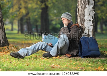 A thoughtful young student seated on a green grass reading a book in the park