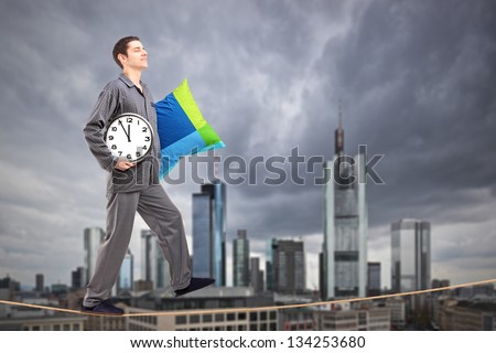 A man in pajamas holding a pillow and clock sleepwalking on a rope, with the financial centre in Frankfurt, Germany in the background