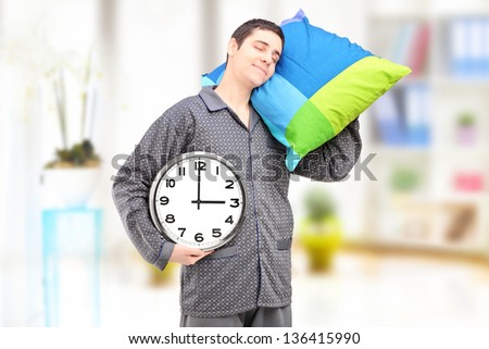 A young lazy guy holding a clock and sleeping on a pillow at home
