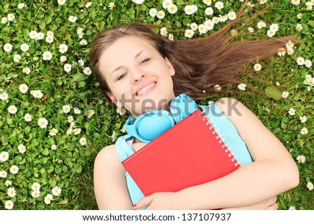A young female with notebook listening music and lying on a grass with daisies