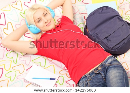 Female student lying in bed with backpack and notebook and listening to music