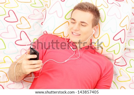 Young smiling man lying on a bed and listening music from his mobile phone