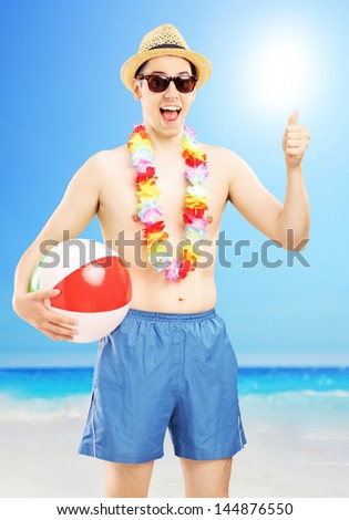 Smiling male in swimming shorts, holding a beach ball and giving thumb up, on a beach next to a sea