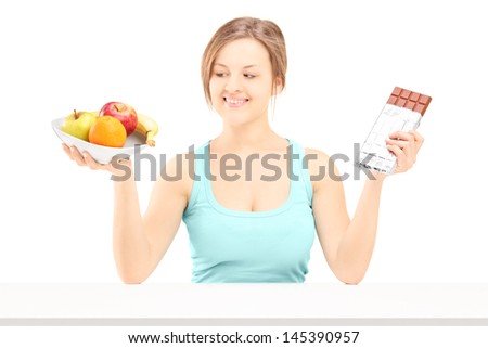 Young female holding a bowl of fresh fruit and chocolate, trying to decide which one to eat, isolated on white background