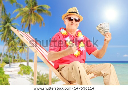 Smiling gentleman sitting on a beach chair and holding US dollars, on a beach near the sea
