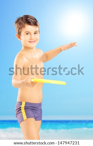 Smiling kid in swimming shorts throwing frisbee on a beach next to the sea