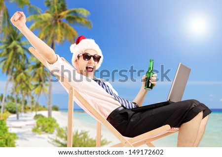 Excited man with santa hat on a beach chair holding a beer and working on a laptop, on a tropical beach