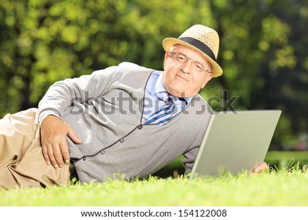 Senior man with hat lying on a green grass and working on a laptop in a park