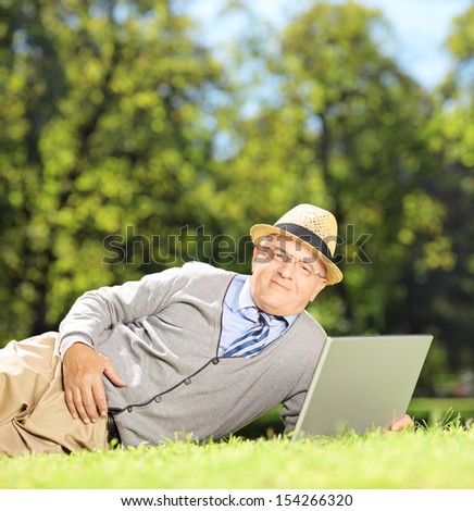 Senior man with hat lying on a green grass and working on a laptop in a park, shot with a tilt and shift lens