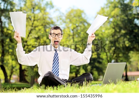Worried businessman holding documents and shouting seated on a green grass in a park