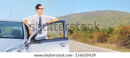 Smiling guy with glasses posing next to his automobile on an open road, shot with a tilt and shift lens