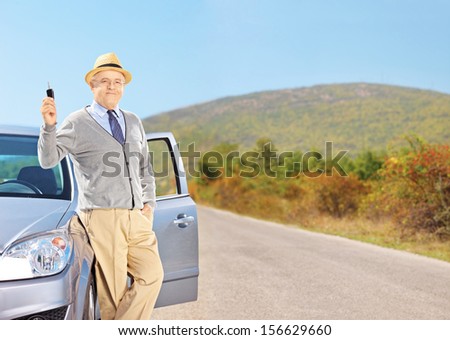 Smiling senior male holding a car key next to his automobile on an open road, shot with a tilt and shift lens