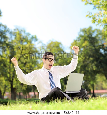 Young man cheering and watching TV on a laptop in a park on a sunny day, shot with a tilt and shift lens