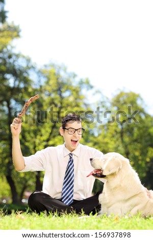 Guy with tie and glasses seated on a green grass playing with labrador retriver in a park