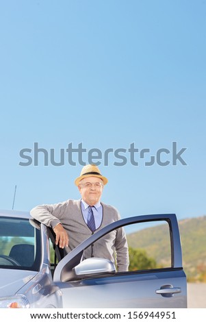 Confident mature gentleman with hat posing next to his automobile on an open road