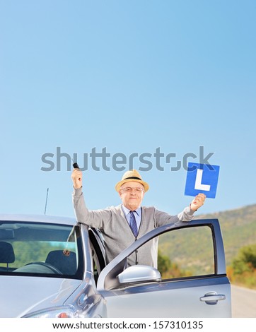 Smiling mature man posing next to his car holding a L sign and car key after having his driver\'s licence, outside, shot with a tilt and shift lens