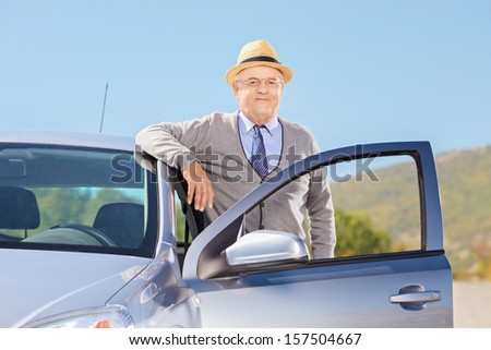 Smiling mature gentleman with hat posing next to his car on an open road