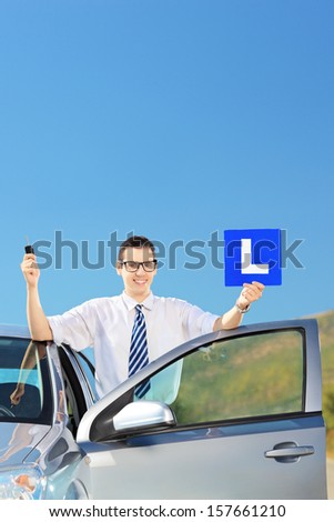 Happy man posing near his car, holding a L sign and car key after having his driver\'s licence on a road