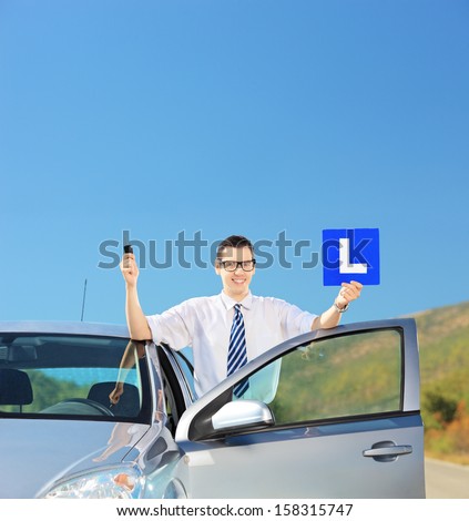 Happy man posing near his car, holding a L sign and car key after having his driver\'s licence on an open road, shot with a tilt and shift lens