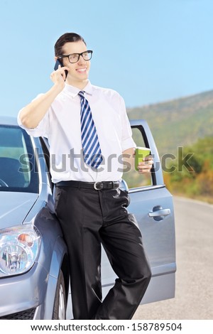 Smiling young man on his automobile drinking coffee and talking on a mobile phone, on an open road