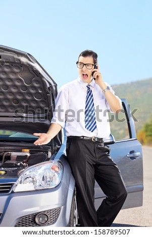 Young nervous man standing next to his broken car and talking on a cell phone, on a sunny day