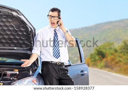 Young nervous man standing next to his broken car and talking on a mobile phone, on a sunny day