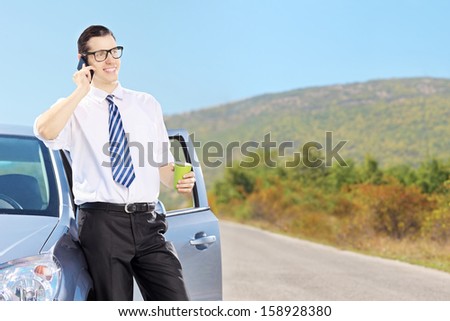 Smiling young man on his automobile talking on a mobile phone and drinking coffee, on an open road