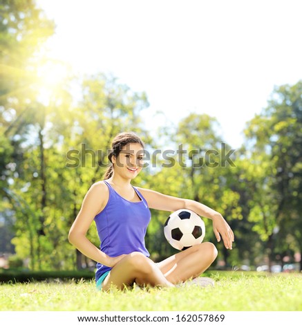 Young athlete female sitting on a green grass and holding a soccer ball in a park, shot with a tilt and shift lens