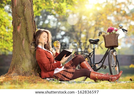 Young female seated on a grass in park reading a book and eating apple, on a sunny day
