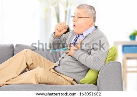 Mature Man Seated On A Sofa Coughing Because Of Pulmonary Disease At Home