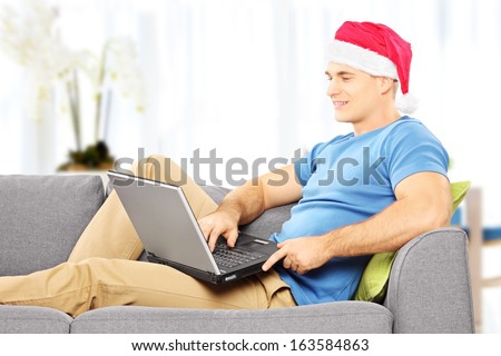 Handsome guy with santa hat seated on a sofa working on a laptop at home