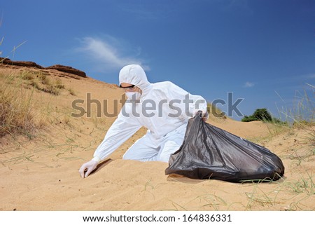 Male worker in protective suit holding a waste bag and collecting samples from sand, symbolizing pollution