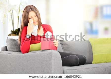 Sad young female seated on a sofa wiping her eyes from crying with tissue at home