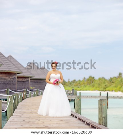 Bride in wedding dress holding a bouquet flowers and posing near the water villas on a Maldives island