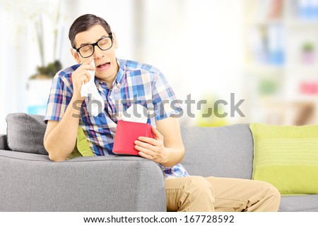 Sad male seated on a sofa wiping his eyes from crying at home