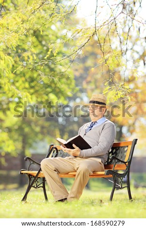 Senior man with hat seated on a wooden bench reading a book outside