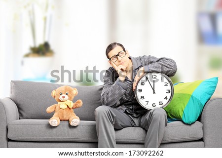 Young man in pajamas holding a big wall clock seated on couch with teddy bear next to him, at home
