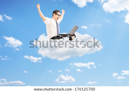 Young businessman working on a laptop and gesturing happiness seated on cloud with blue sky in the background