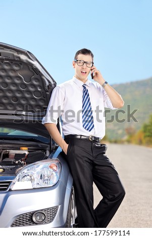 Male businessperson talking on phone next to a car with its bonnet open on an open road.